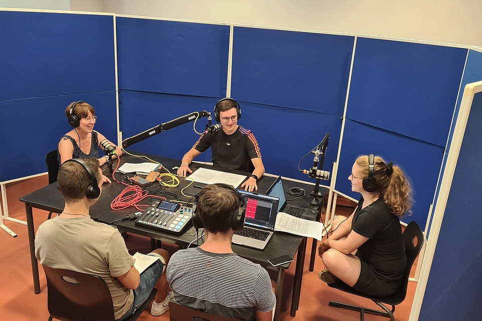 Beispielbild Podcastaufnahme: 5 Personen, 3 Sprecher mit Mikrofonen und 2 Menschen mit Aufnahmepult 
